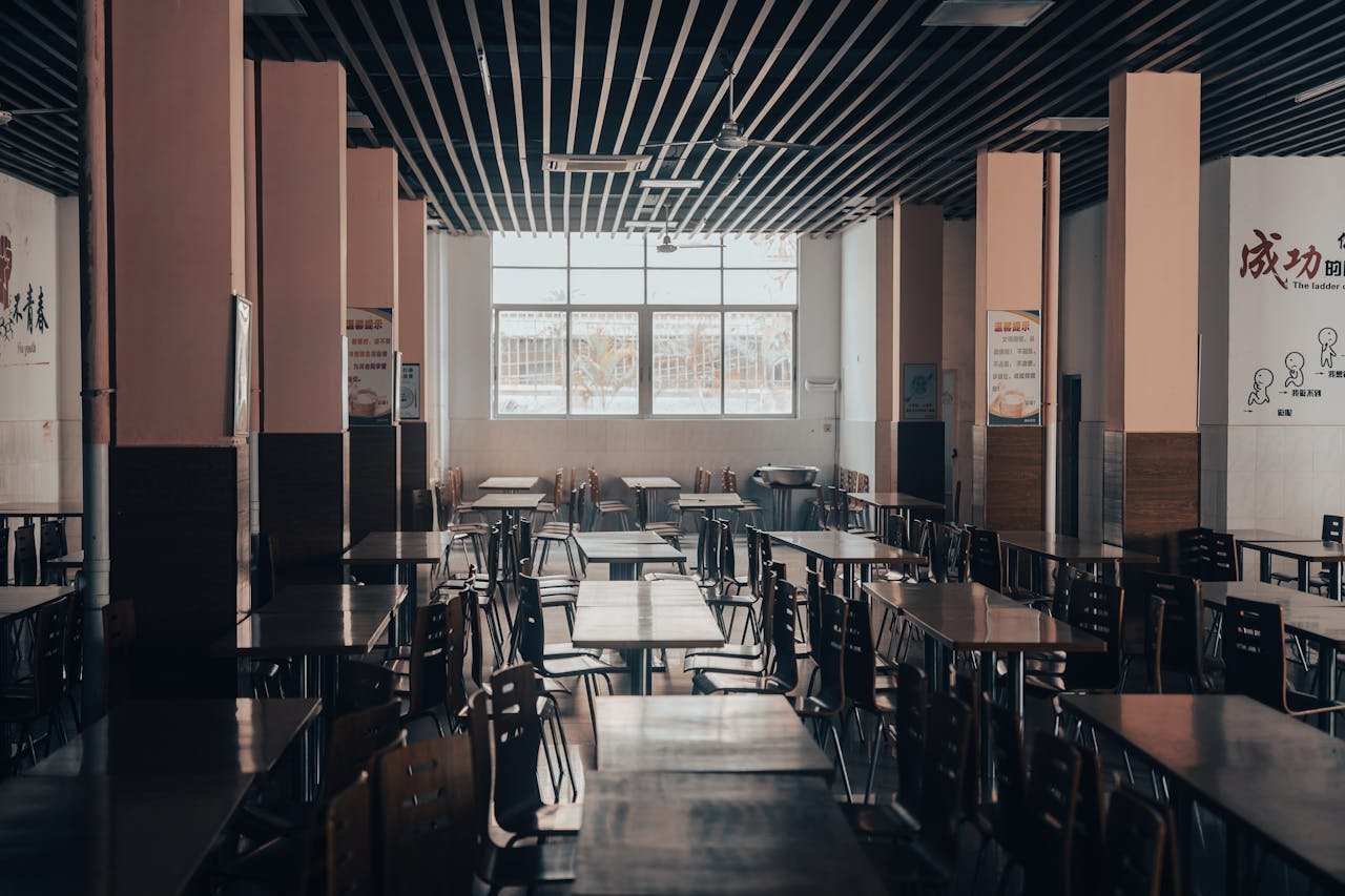 An empty cafeteria featuring wooden tables and chairs, lit by natural light through large windows.